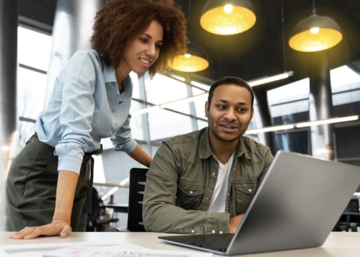 Uma mulher jovem, vestindo camisa azul-clara, e um homem sentado, vestindo jaqueta verde, analisam e discutem juntos o conteúdo na tela de um notebook sobre uma mesa de escritório.